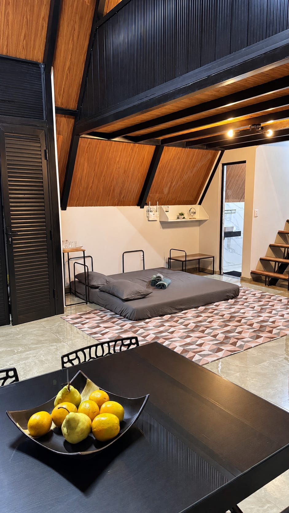 Mesa de refeições preta com fruteira em primeiro plano e, ao fundo, o sofá-cama aberto sobre o tapete e a escada para o mezanino.