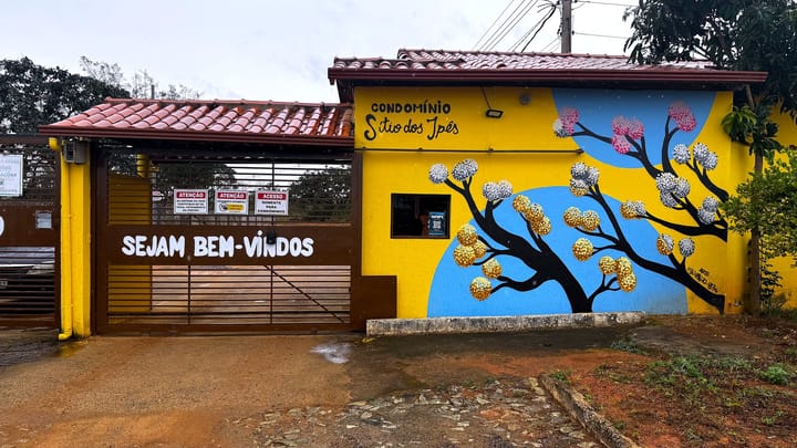 A portaria do condomínio com o portão, a faixa de boas-vindas e o mural de ipês na parede amarela.