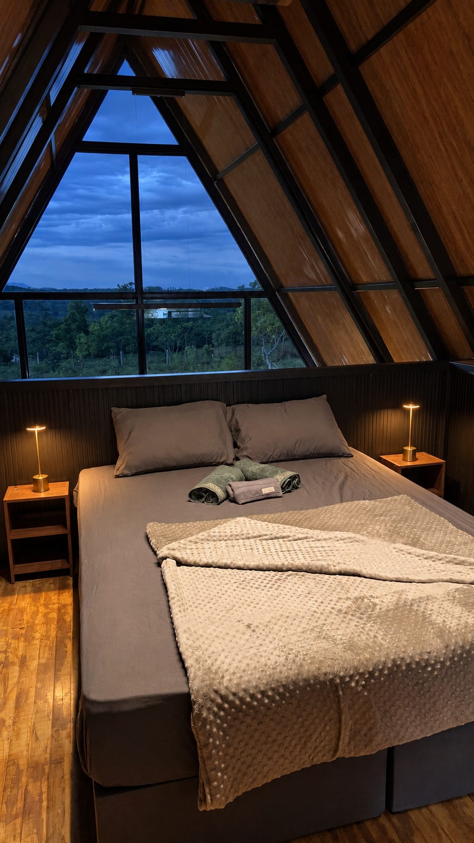Cama de casal com manta clara e dois abajures acesos, de frente para a janela triangular, com o cerrado e o céu do anoitecer lá fora.