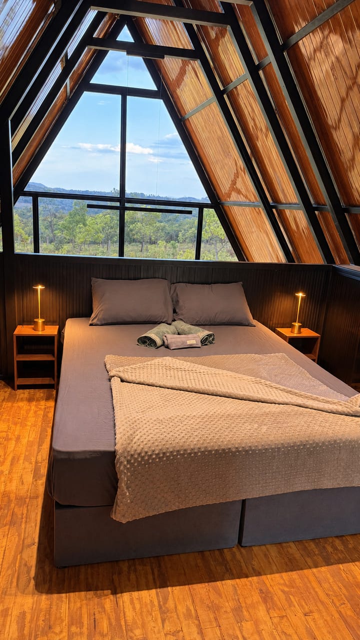 A cama queen do mezanino diante da empena de vidro, com o cerrado lá fora.
