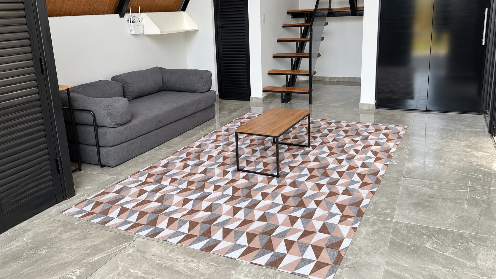 A sala com o sofá cinza, a mesa de centro de madeira, o tapete geométrico e a escada ao fundo.