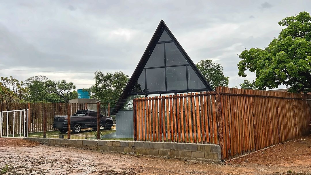 Chalé A-frame com a empena de vidro em triângulo, atrás de uma cerca de ripas de madeira, em dia de chuva.