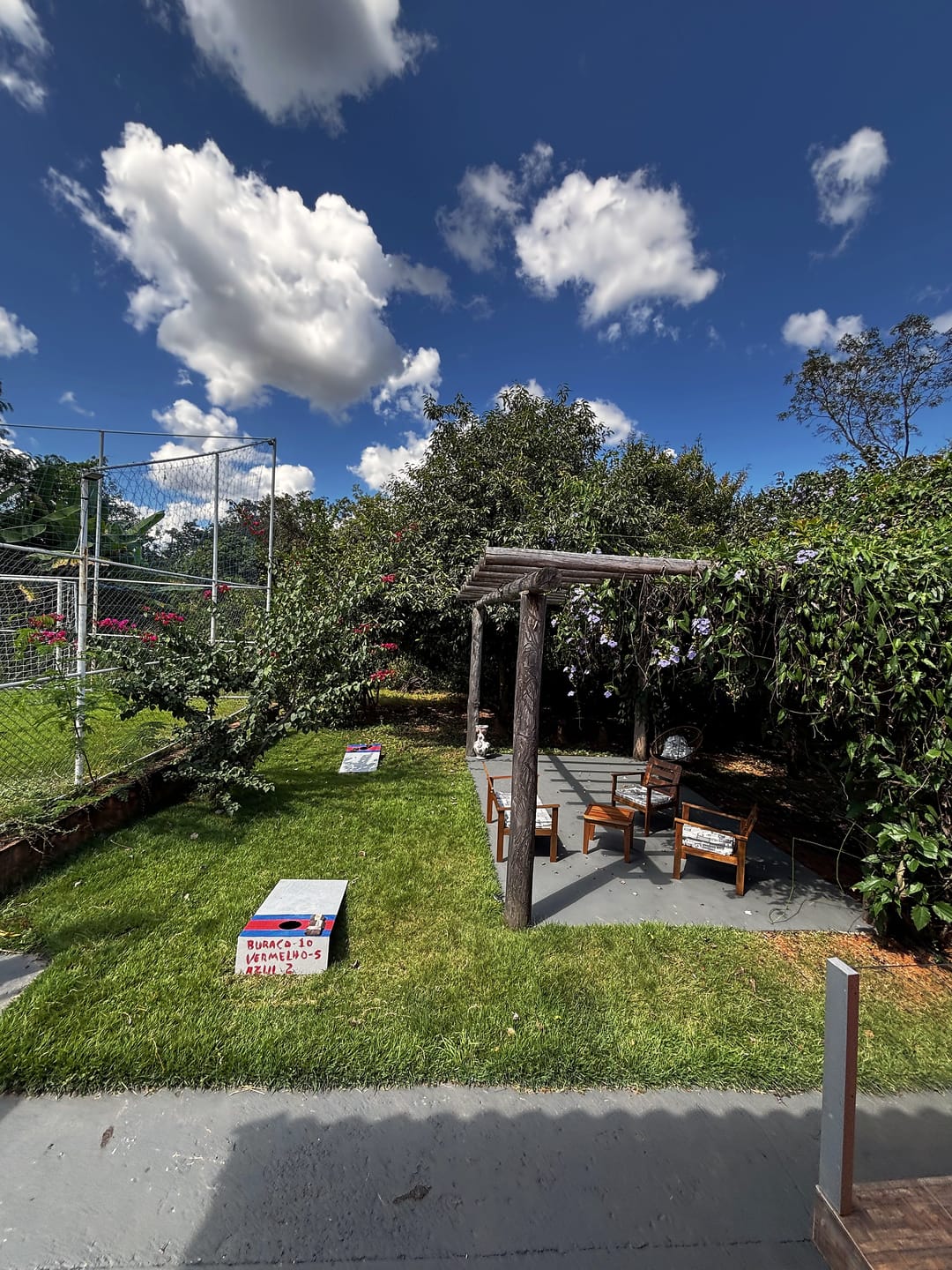 Gramado com pergolado de madeira, cadeiras e jogos ao ar livre, sob céu azul com nuvens.