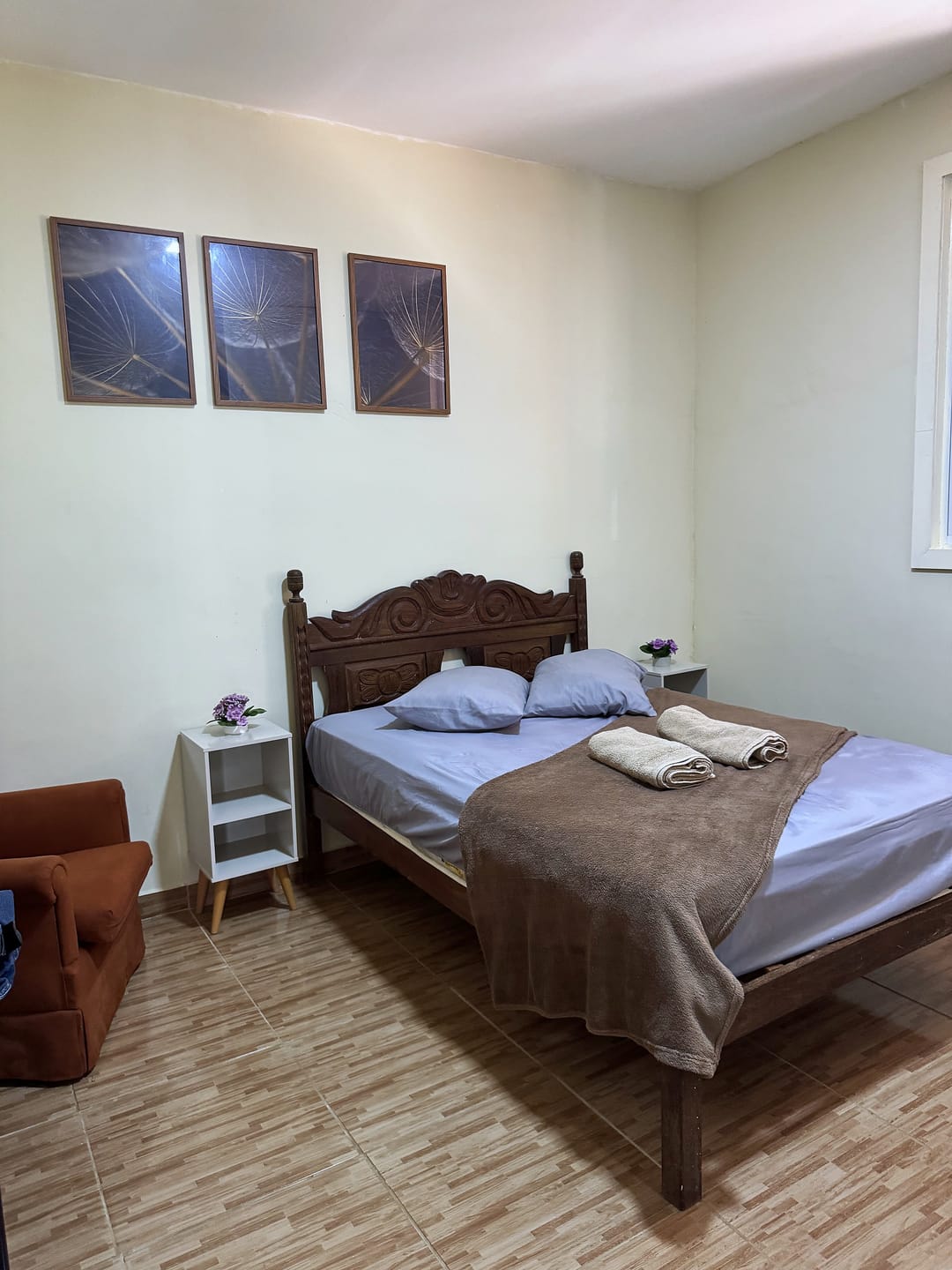 Quarto com cama de casal de madeira entalhada, toalhas dobradas sobre a cama e três quadros na parede.