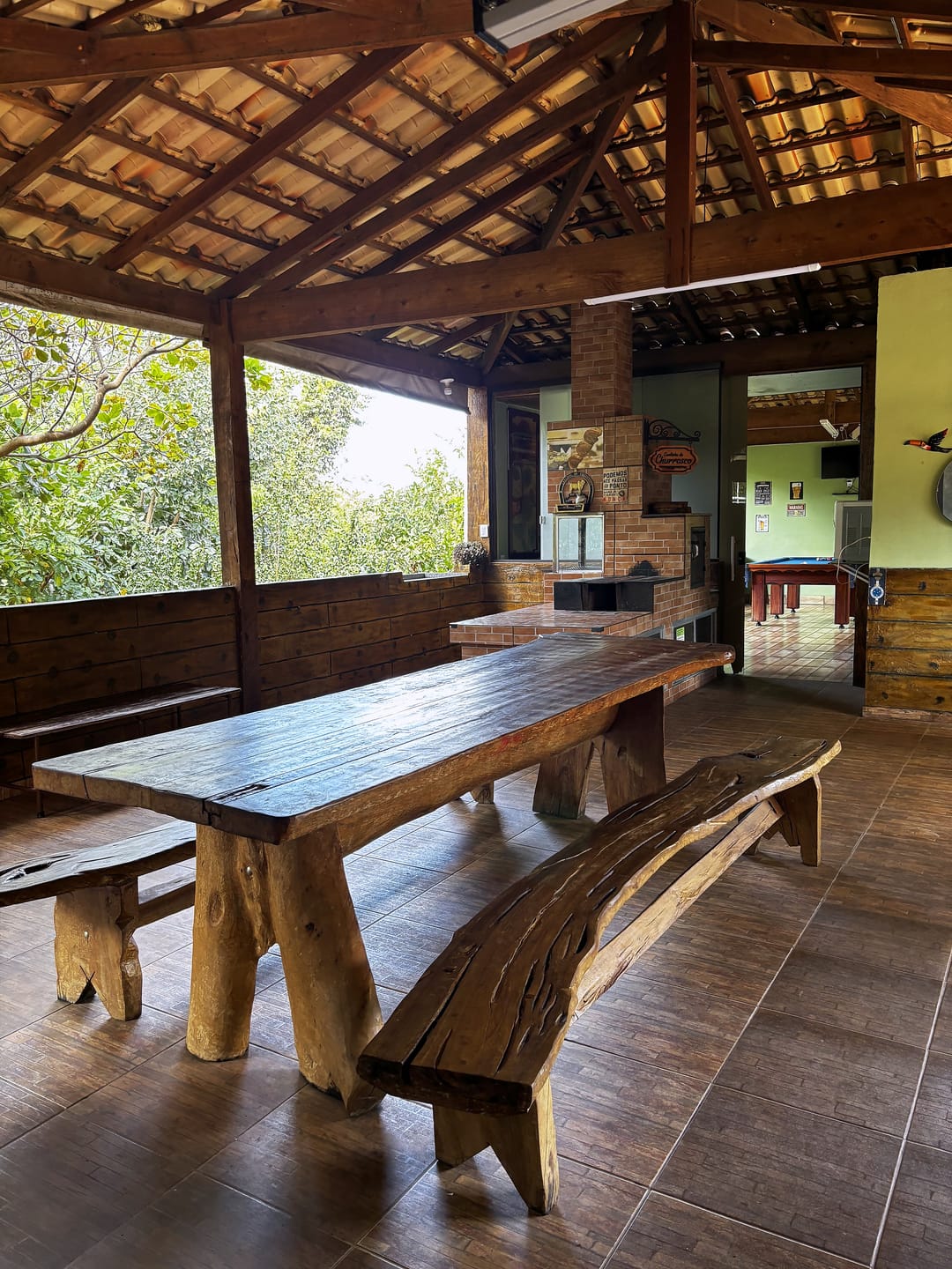 Mesa longa de madeira rústica, com bancos, sob o telhado de madeira do rancho coberto.